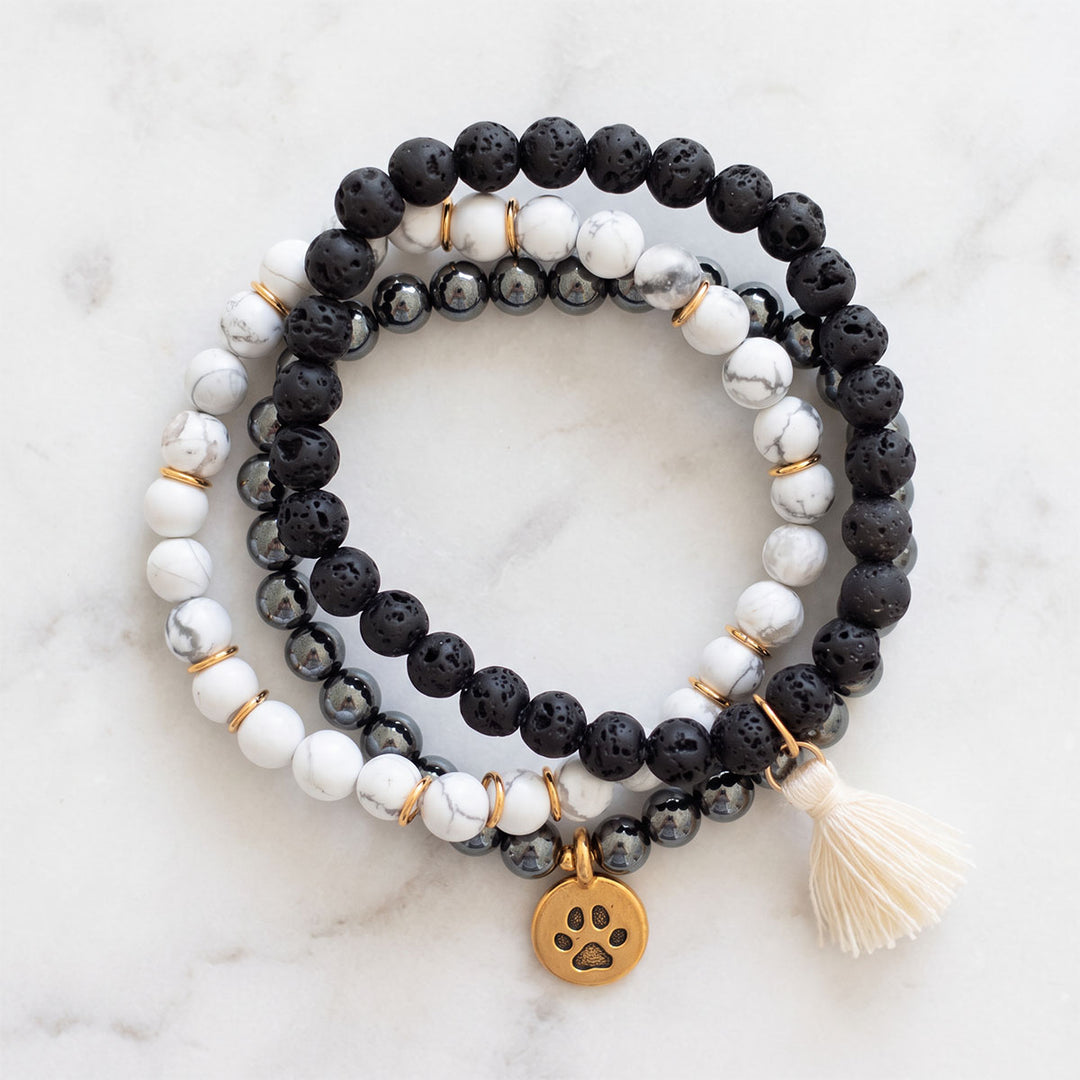 Three coordinating gemstone bracelets arranged together on a pale marble background, featuring polished grey hematite with a gold-plated paw charm, matte black lava stone with a cream tassel, and white howlite with gold-plated accents.