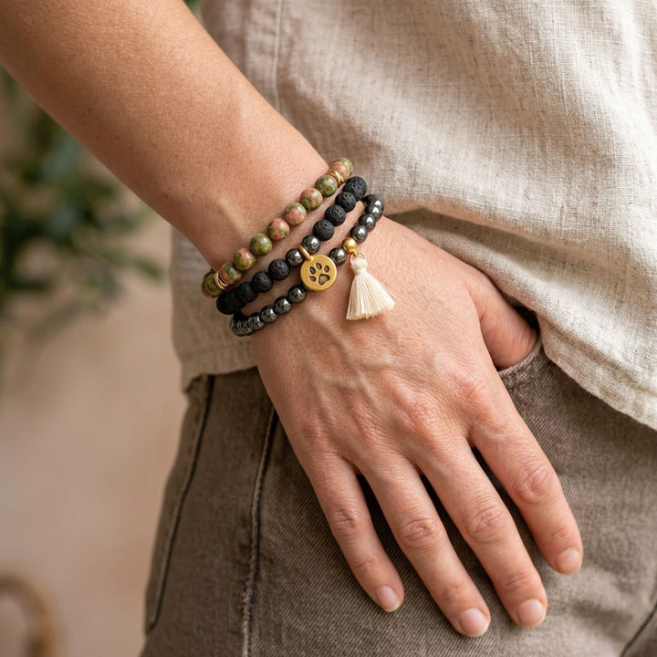 Three gemstone bracelets worn together on a wrist with neutral linen clothing, showing green and pink unakite, black lava stone, polished hematite, a gold-plated paw charm and a cream tassel.