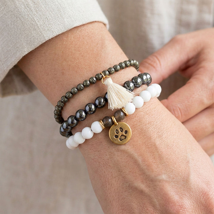 Close-up of three gemstone bracelets worn stacked on a wrist model, showing white jade and labradorite with a gold-tone paw charm, polished hematite and fine pyrite beads with a cream tassel.