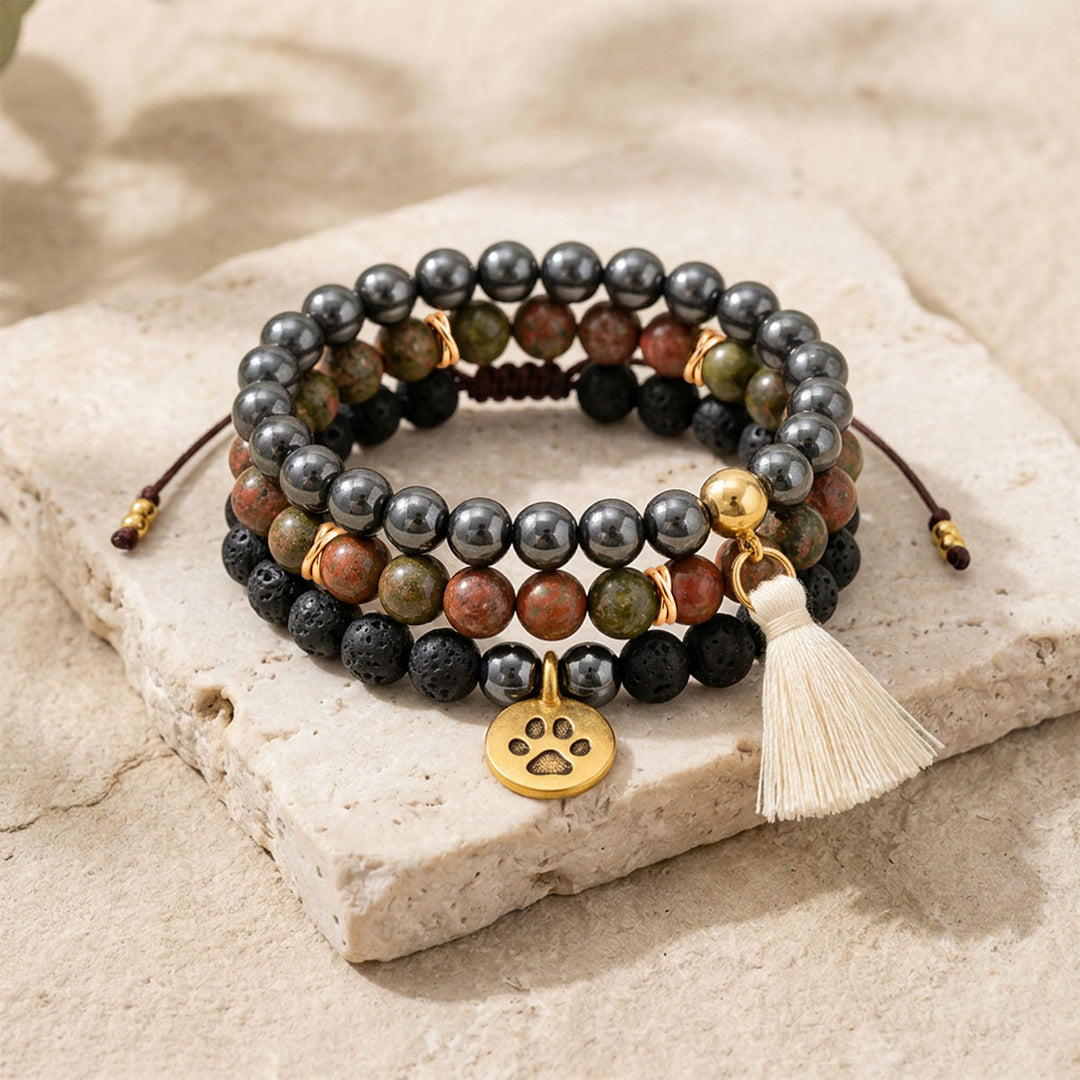 Close-up of a three-bracelet gemstone stack arranged on pale stone, featuring black lava beads, metallic hematite, green and pink unakite, a gold-plated paw charm and a cream cotton tassel.