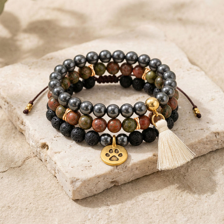 Close-up of a three-bracelet gemstone stack arranged on pale stone, featuring black lava beads, metallic hematite, green and pink unakite, a gold-plated paw charm and a cream cotton tassel.