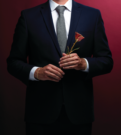 A handsome man is shown in a navy blue suit. He's holding a red Everlasting Rose in his hands.