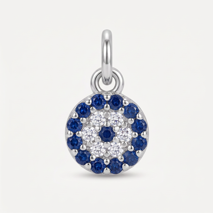 A round sterling silver charm pendant featuring a multi-layered evil eye design with blue sapphires and clear cubic zirconia stones, shown on a light gray background.