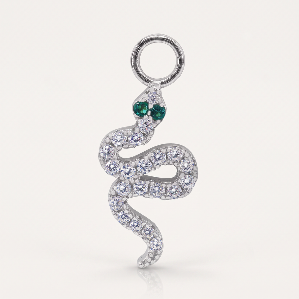 A silver snake pendant adorned with clear pavé stones and featuring two green stone eyes, shown on a light cream background.