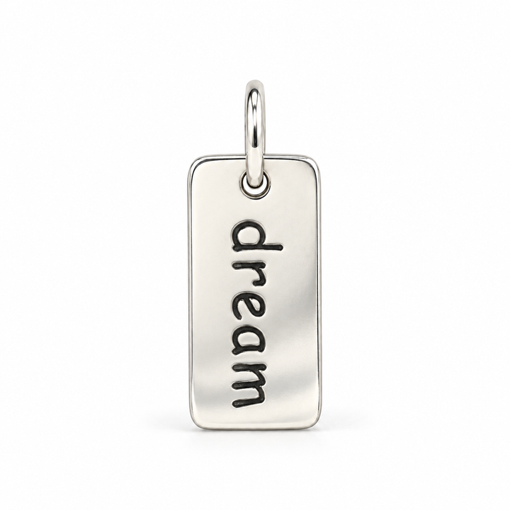 Rectangular sterling silver tag charm with rounded corners, engraved with “dream” in lowercase letters, with a silver jump ring.